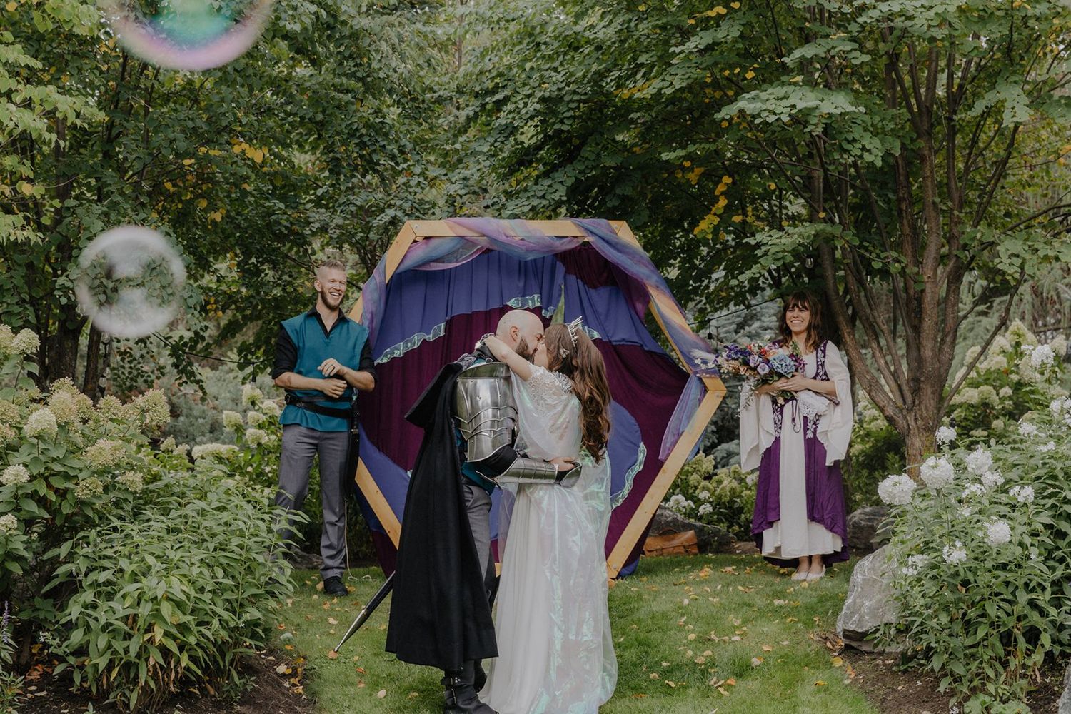 Couple sharing a kiss under a decorative arch at a fantasy-themed outdoor wedding ceremony.