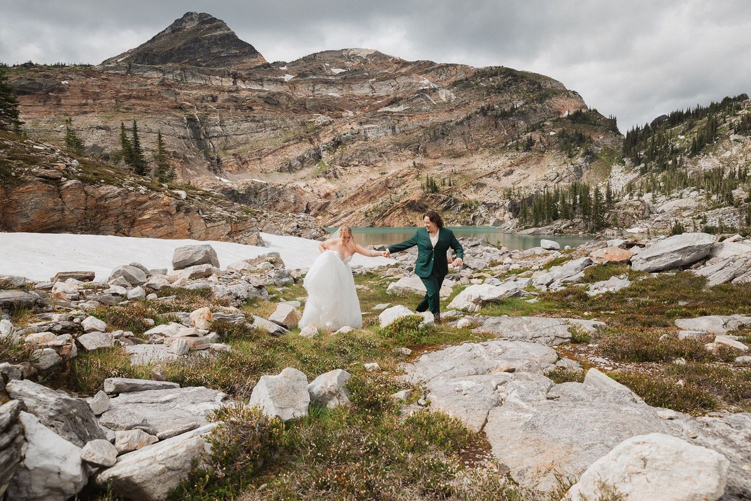Two brides walk hand-in-hand through a rocky alpine meadow with mountain peaks behind.