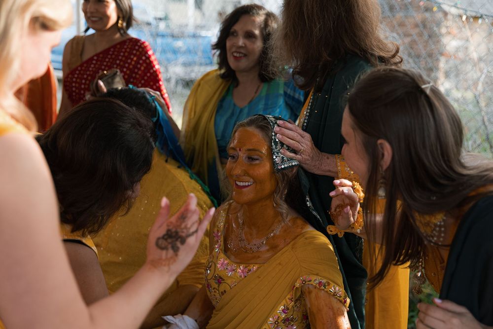 Bride in yellow sari laughs with friends during joyful Indian pre-wedding haldi ceremony.