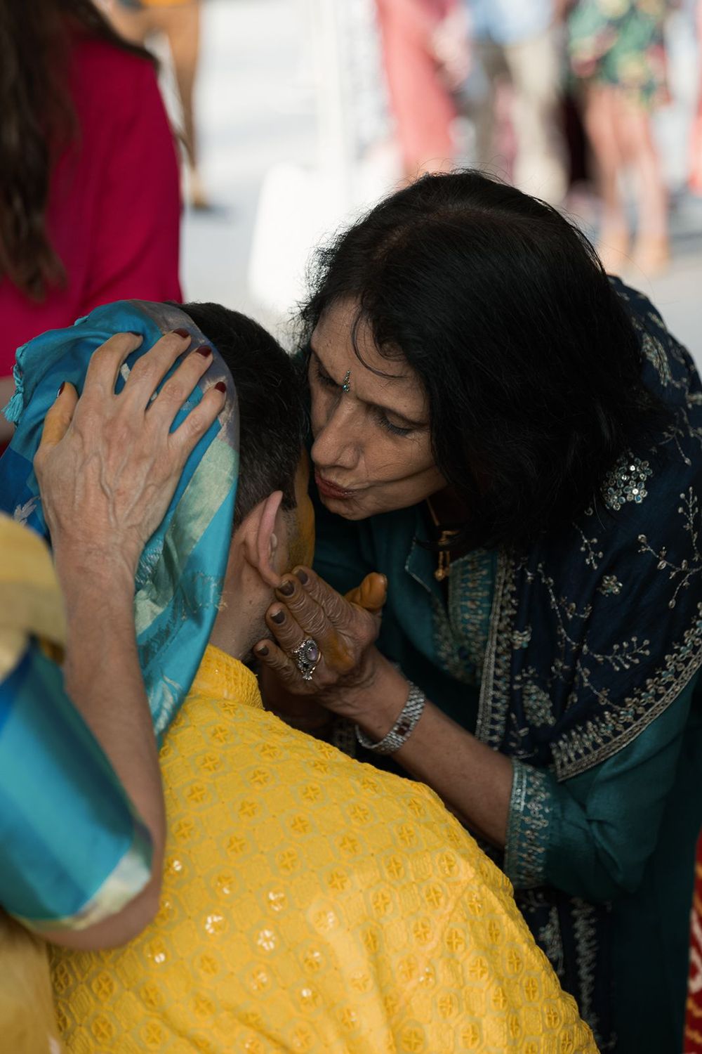 Emotional Indian woman in teal sari comforting a young woman in yellow lehenga at a wedding celebration.