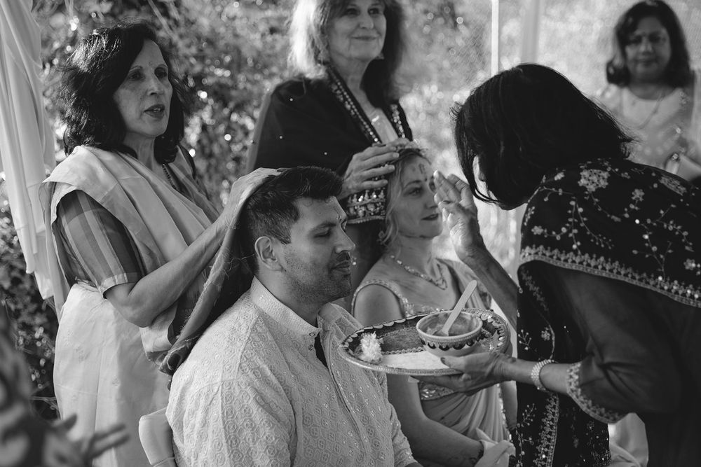 Black and white photo of guests participating in traditional Indian wedding ceremony rituals indoors.