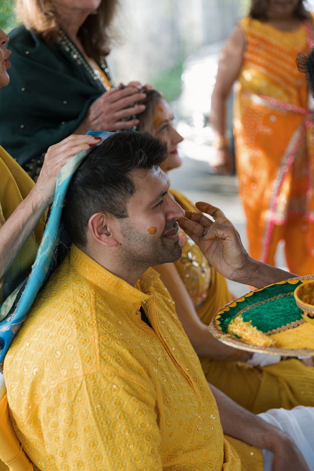 Man in yellow kurta receives haldi turmeric paste application on face during pre-wedding ceremony.