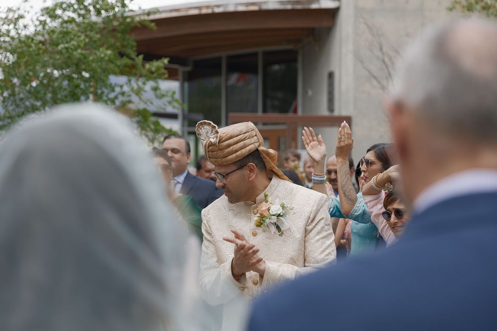 Indian groom in cream sherwani and turban walks through celebrating wedding guests at outdoor ceremony.