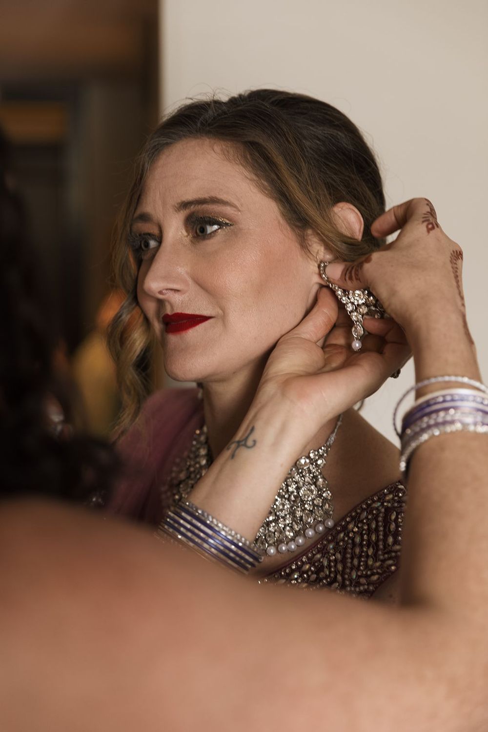 Bride in beaded gown with red lips has ornate chandelier earring placed by assistant before wedding.