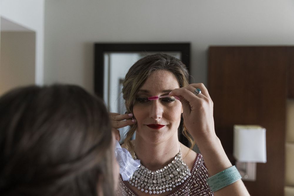 Bride with bold red lips applies mascara while looking in mirror wearing beaded wedding attire.