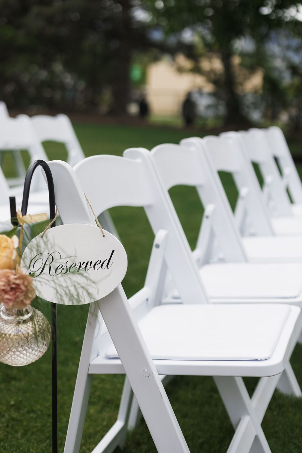 White folding chairs with Reserved sign and floral arrangement set up for outdoor wedding ceremony.
