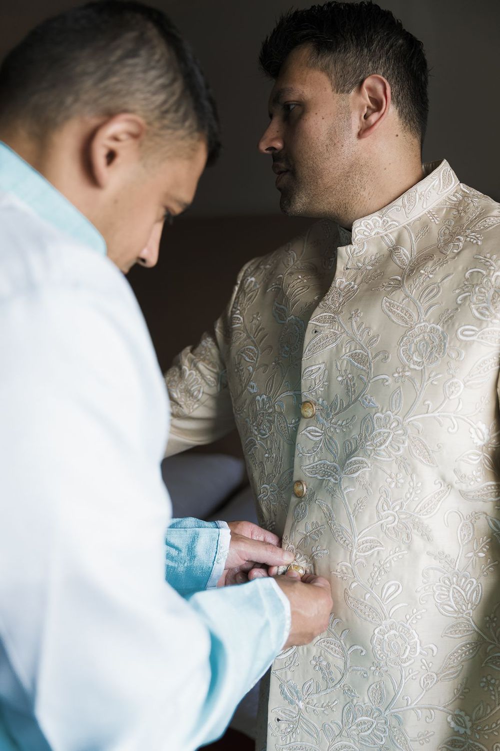 Two men in traditional Indian wedding attire helping each other get dressed indoors.