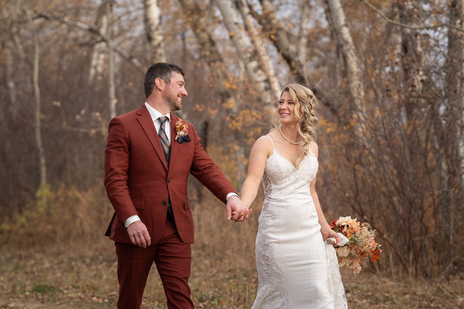 A couple in wedding attire walks down a wooded autumn path together in a romantic outdoor setting.