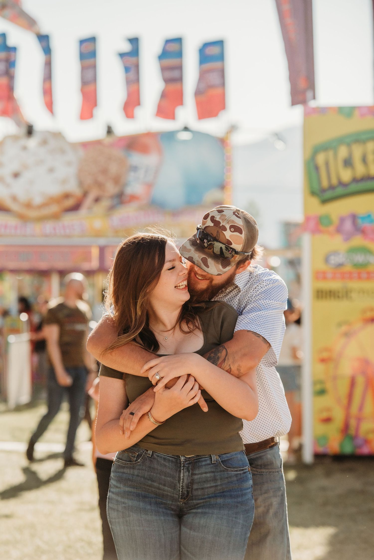 A romantic couple embraces and shares tender moments at a carnival with colorful vendor stalls in the background.