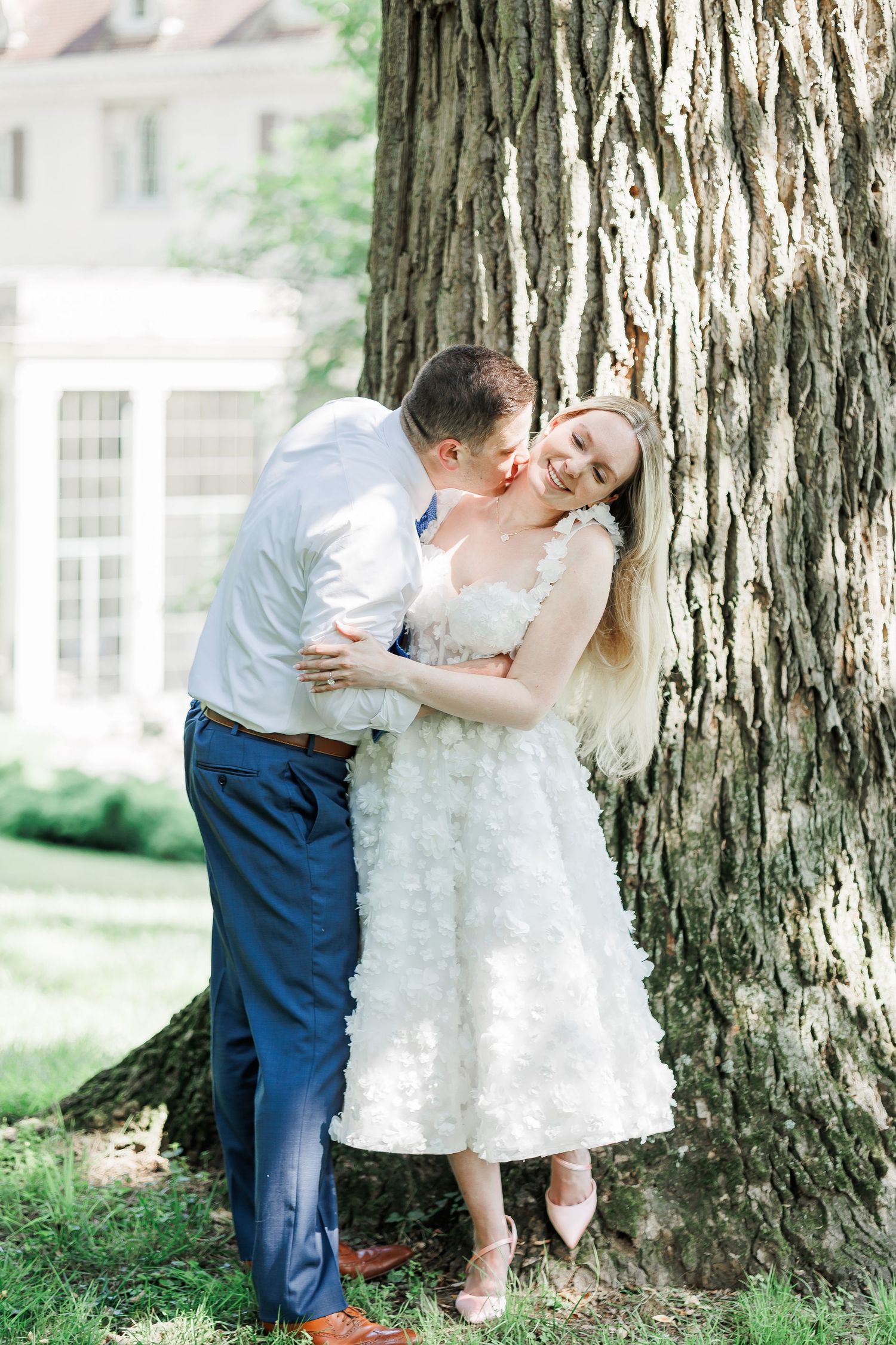 A romantic wedding couple shares a tender moment against a large tree trunk in a sunlit garden setting.