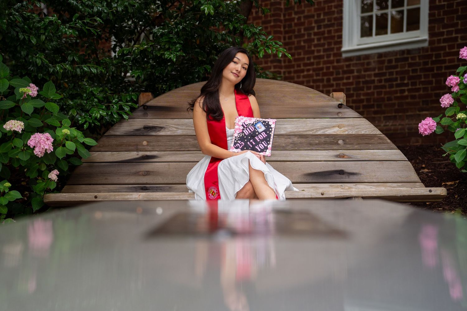 Graduate in white dress and red stole sits on wooden steps surrounded by pink flowers.