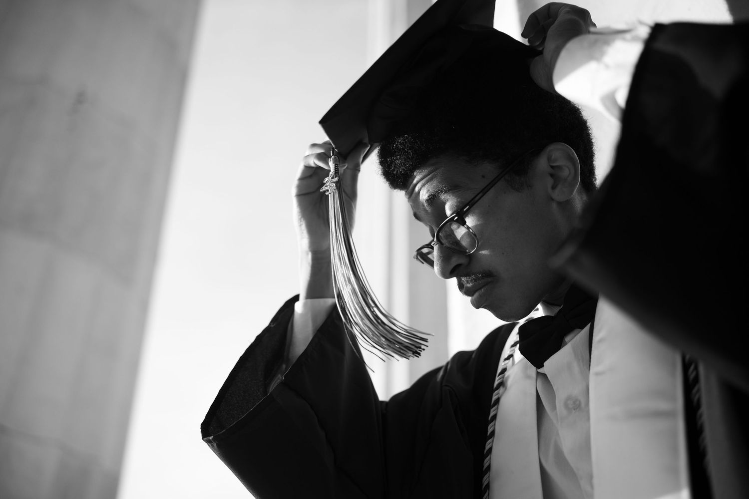 Black and white portrait of graduate adjusting mortarboard cap in dramatic lighting.