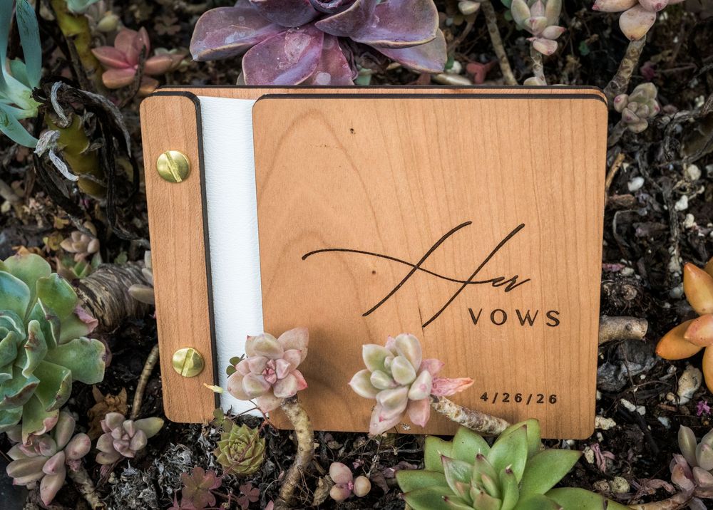 Elegant wooden Her Vows book with engraved script dated 4/26/26 nestled among colorful succulents.