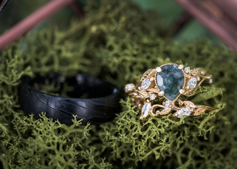Gold ring with teal moss agate and diamond accents on green moss beside a black wedding band.