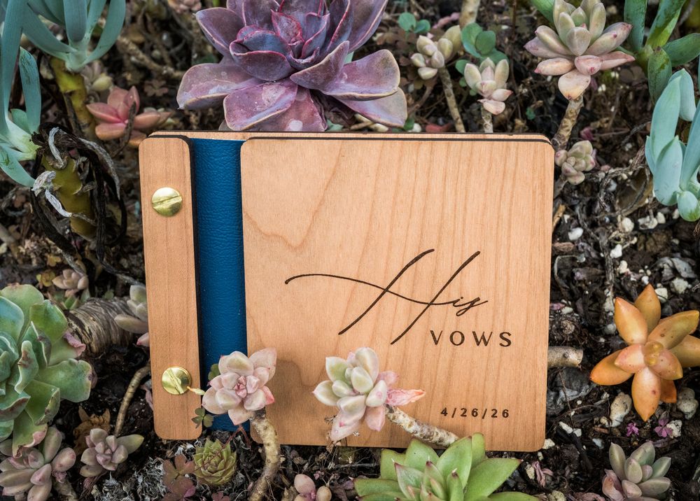 Wooden His Vows keepsake book with blue spine dated 4/26/24 displayed among vibrant succulent plants.
