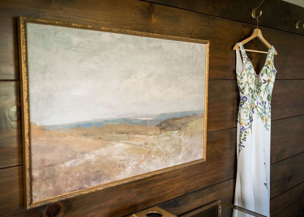 Floral wedding dress hanging beside a vintage landscape painting on a rustic wood-paneled wall.