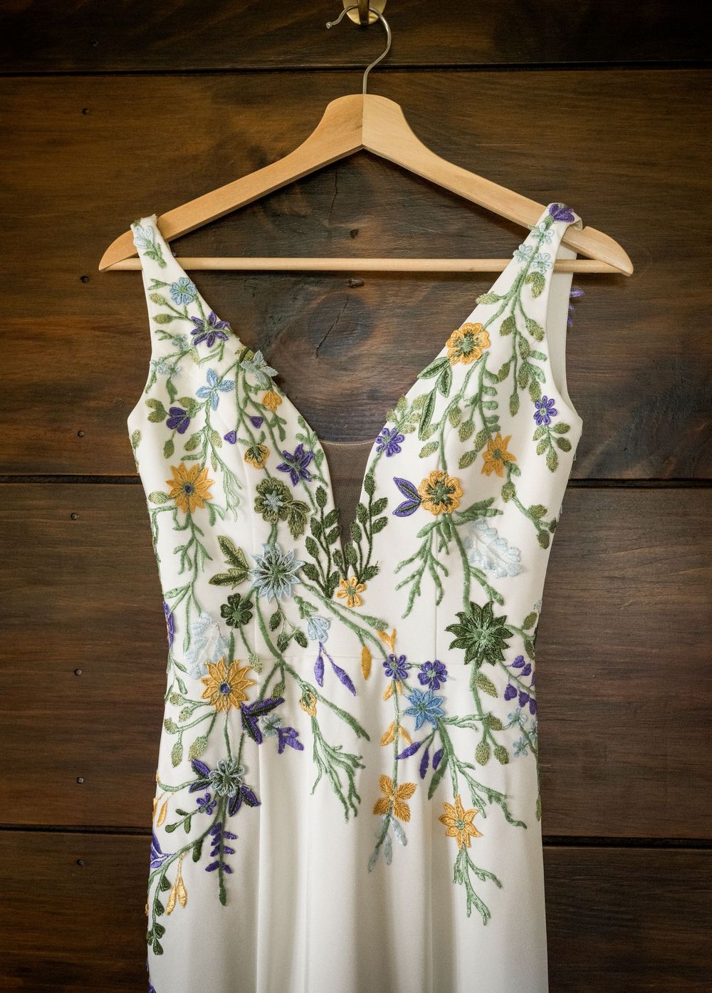 White floral embroidered wedding dress with V-neckline hanging on a wooden hanger against wood wall.