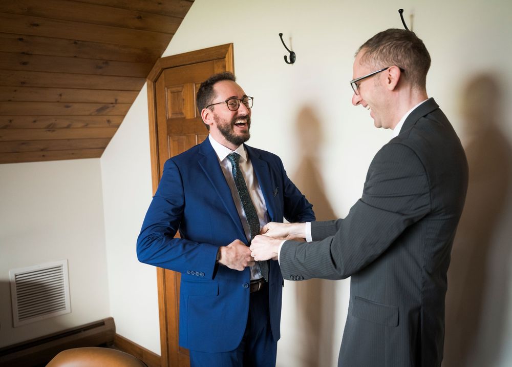Groom in navy suit shakes hands with guest in gray suit inside a rustic wedding venue.