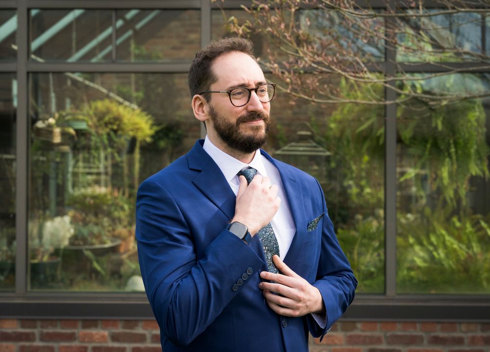 Dapper groom in blue suit adjusts his tie outside a greenhouse before his wedding ceremony.