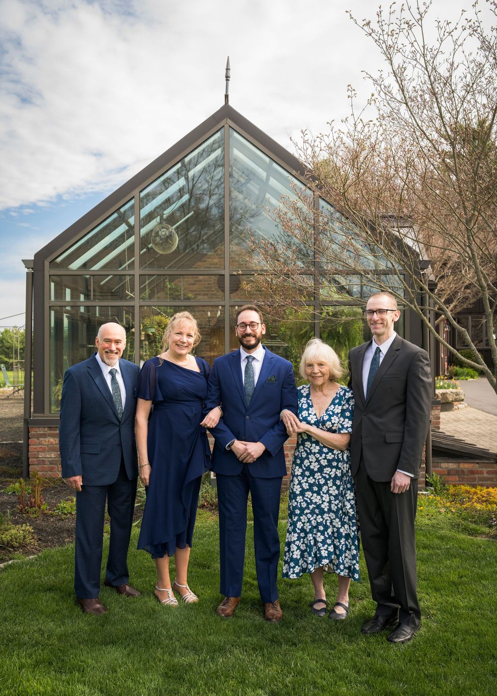 Wedding family portrait of five elegantly dressed guests posing before a glass greenhouse structure.