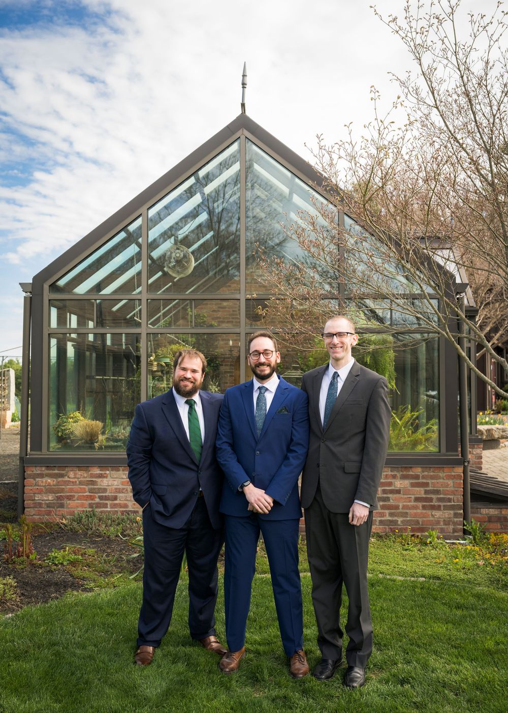 Three groomsmen in suits smiling in front of a glass greenhouse structure on a partly cloudy day.