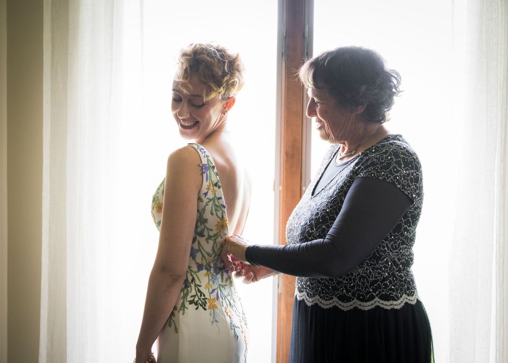 Smiling bride in floral wedding dress being helped by woman near bright window before ceremony.