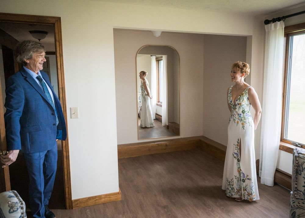Bride in floral gown reflected in mirror while man in blue suit stands nearby.