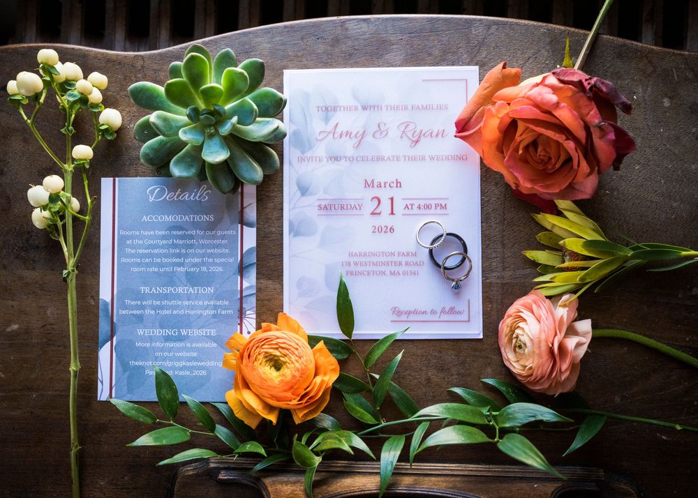 Wedding invitations and stationery arranged with coral roses and greenery on dark wooden surface.