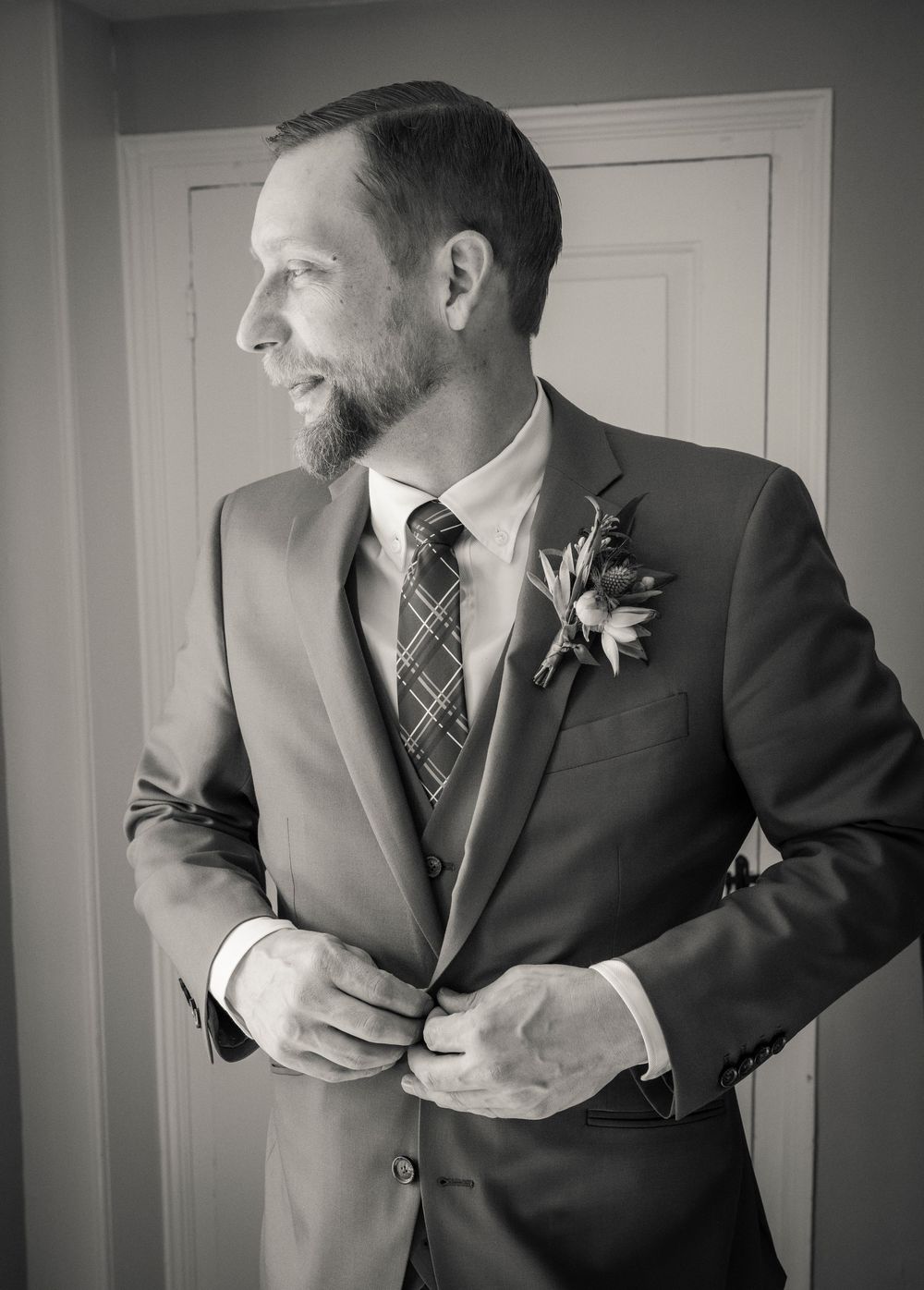 Black and white profile portrait of a groom in a suit and plaid tie with boutonniere standing against a white door.