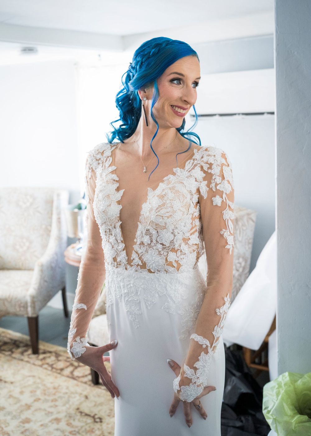 A bride in an elegant lace wedding gown with sheer sleeves poses in a bright indoor setting.