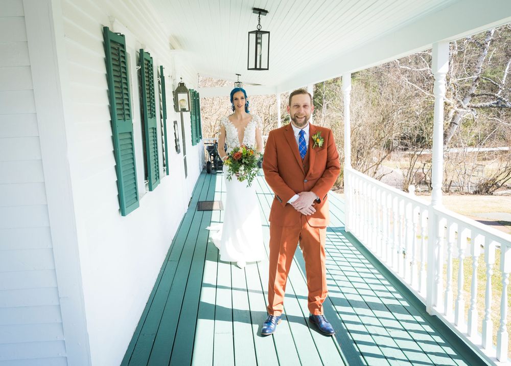 A couple poses on a white porch with green shutters in wedding attire.
