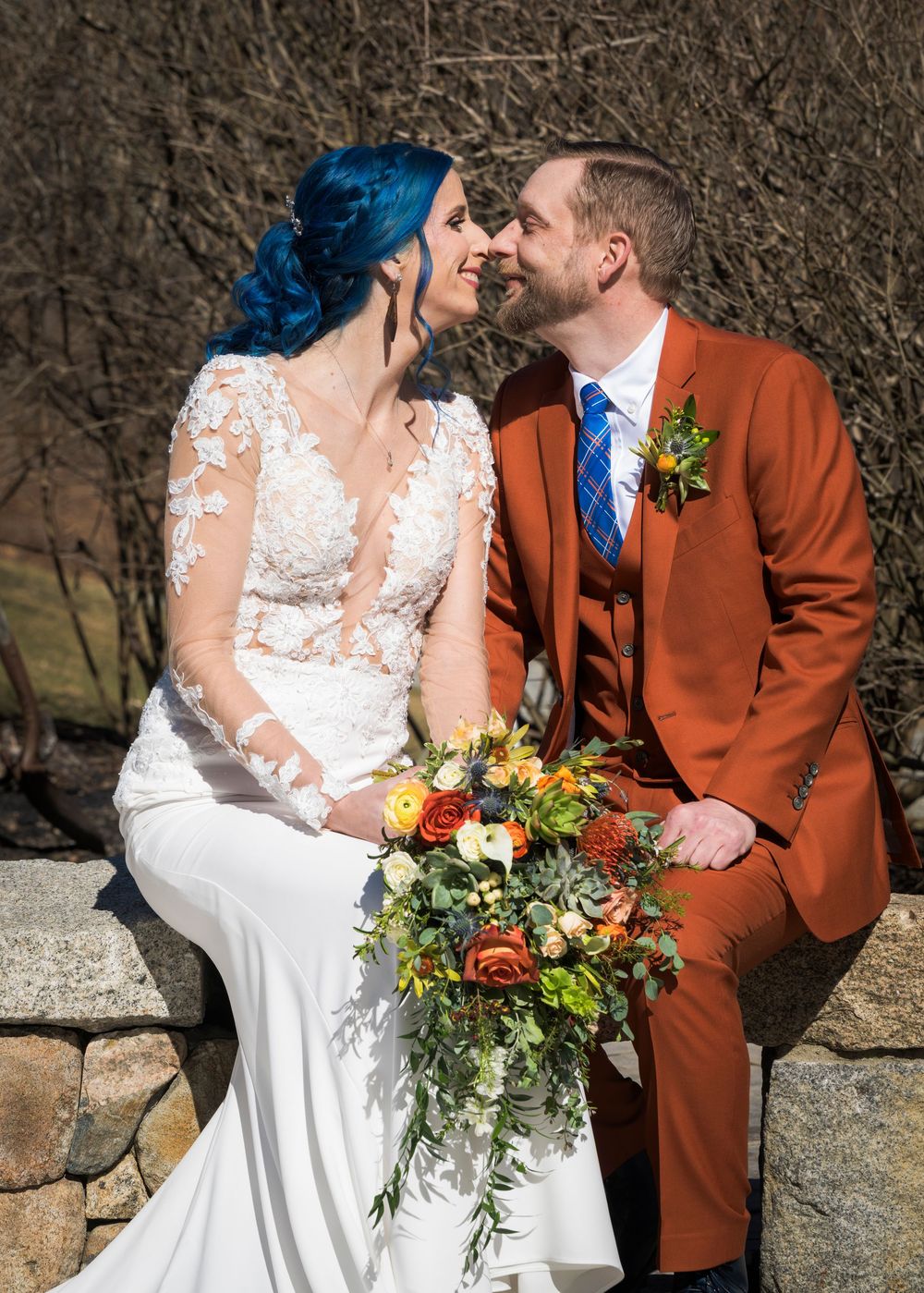 A romantic wedding couple poses against a stone wall backdrop with the bride holding a vibrant autumn bouquet.