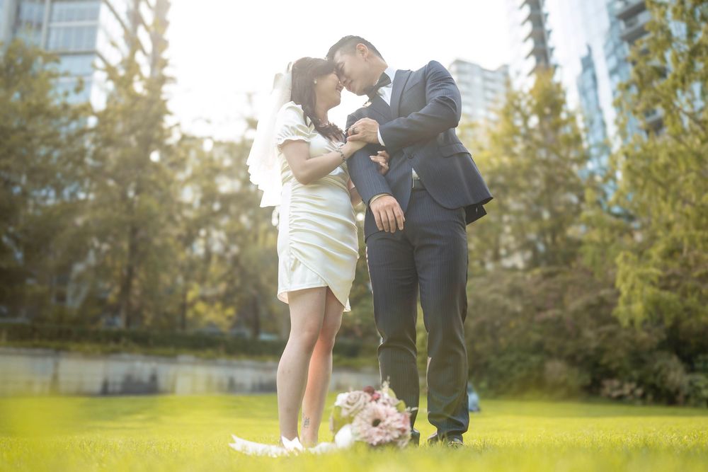 A couple shares a tender moment together in a sunlit park with evergreen trees in the background.