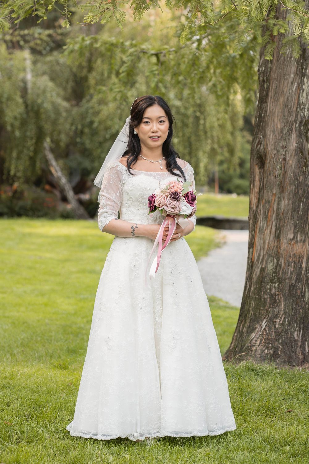 A bride in a white lace dress holds a pink and white bouquet while standing in a park setting.