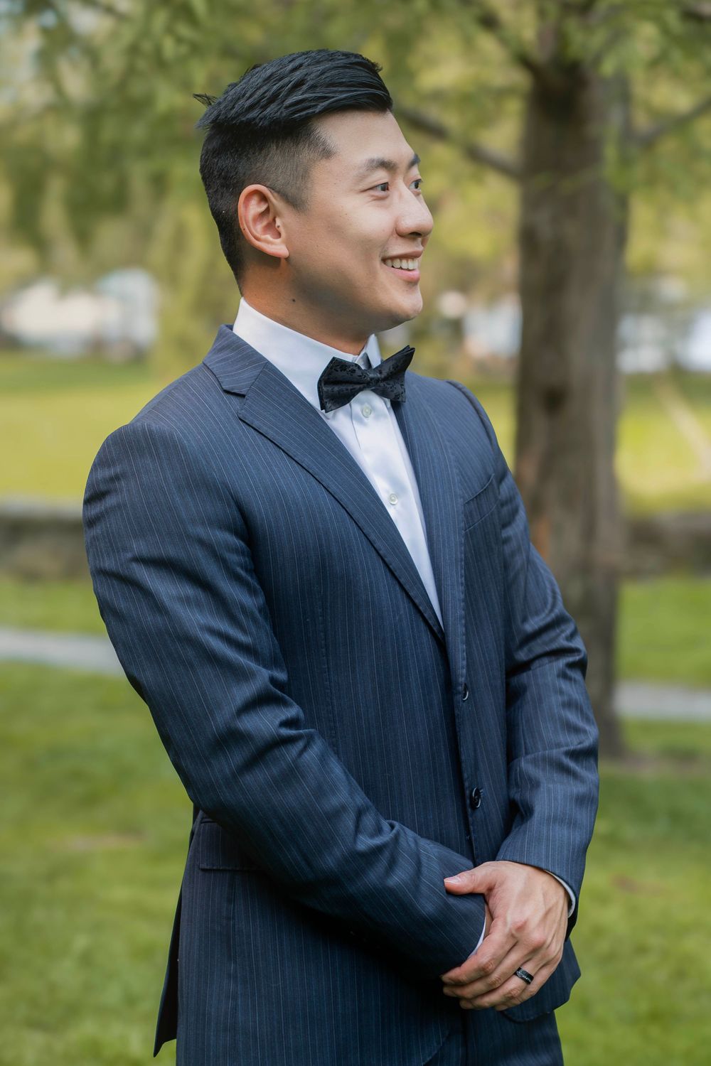 A person in a pinstriped suit and white dress shirt stands outdoors in a park setting with trees in the background.