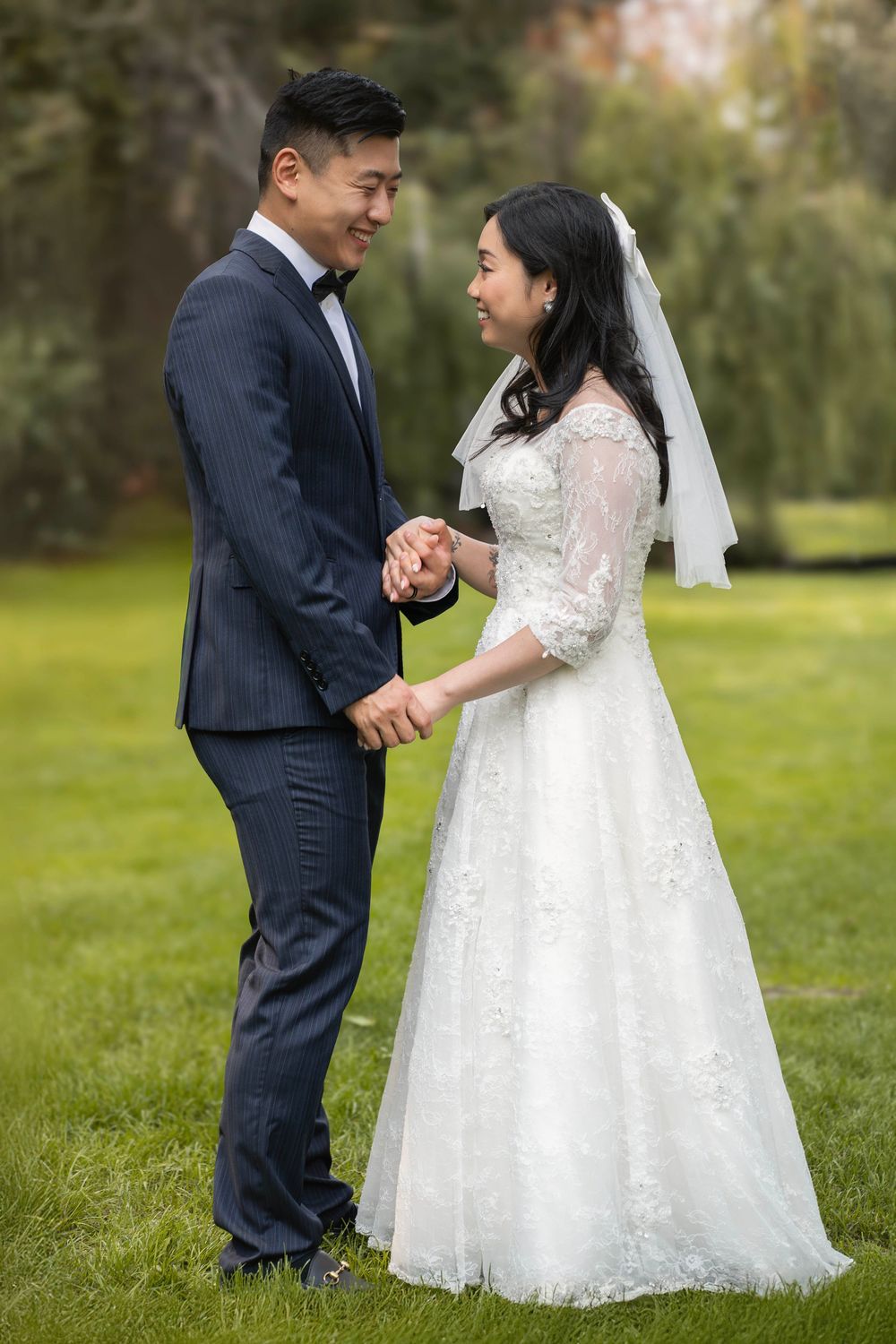 Wedding couple holds hands and gazes at each other on grassy lawn with willows in background.