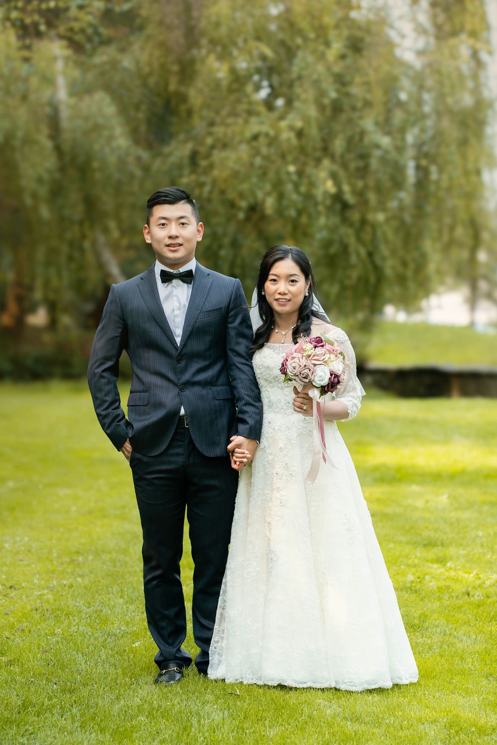 Wedding couple poses formally in front of weeping willow trees holding pink bouquet.