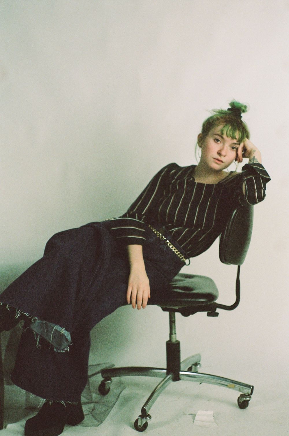 Dramatic editorial poses with green hair on office chair wearing dark pinstriped outfit.