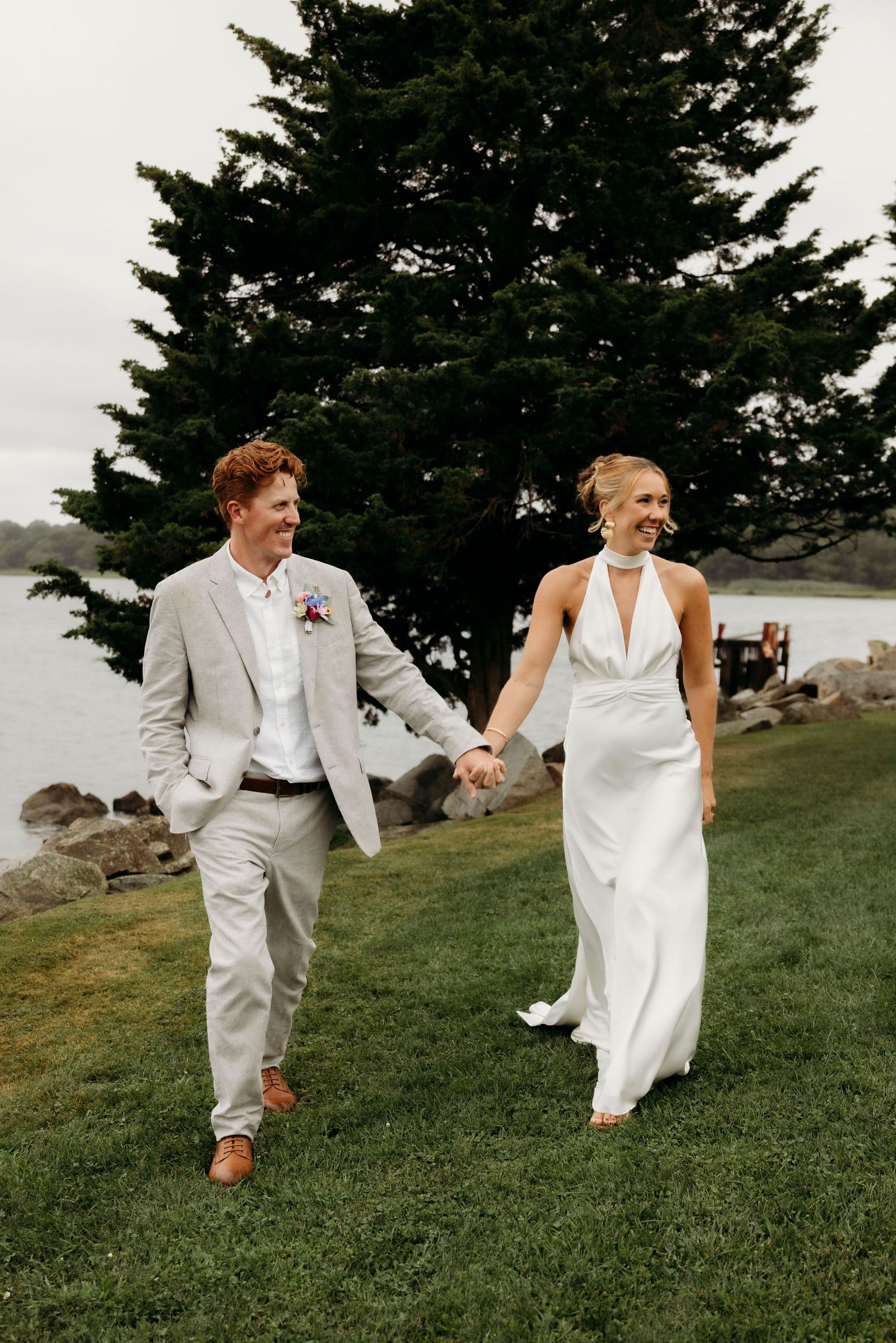 A couple in white wedding attire playfully run and dance together on a grassy hillside with evergreen trees in the background.