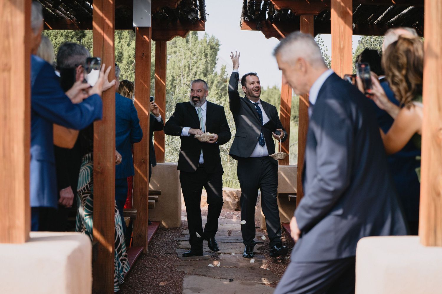 Excited groomsmen celebrate with raised arms as they enter rustic wooden wedding ceremony venue with guests at Blame Her Ranch in Santa Fe