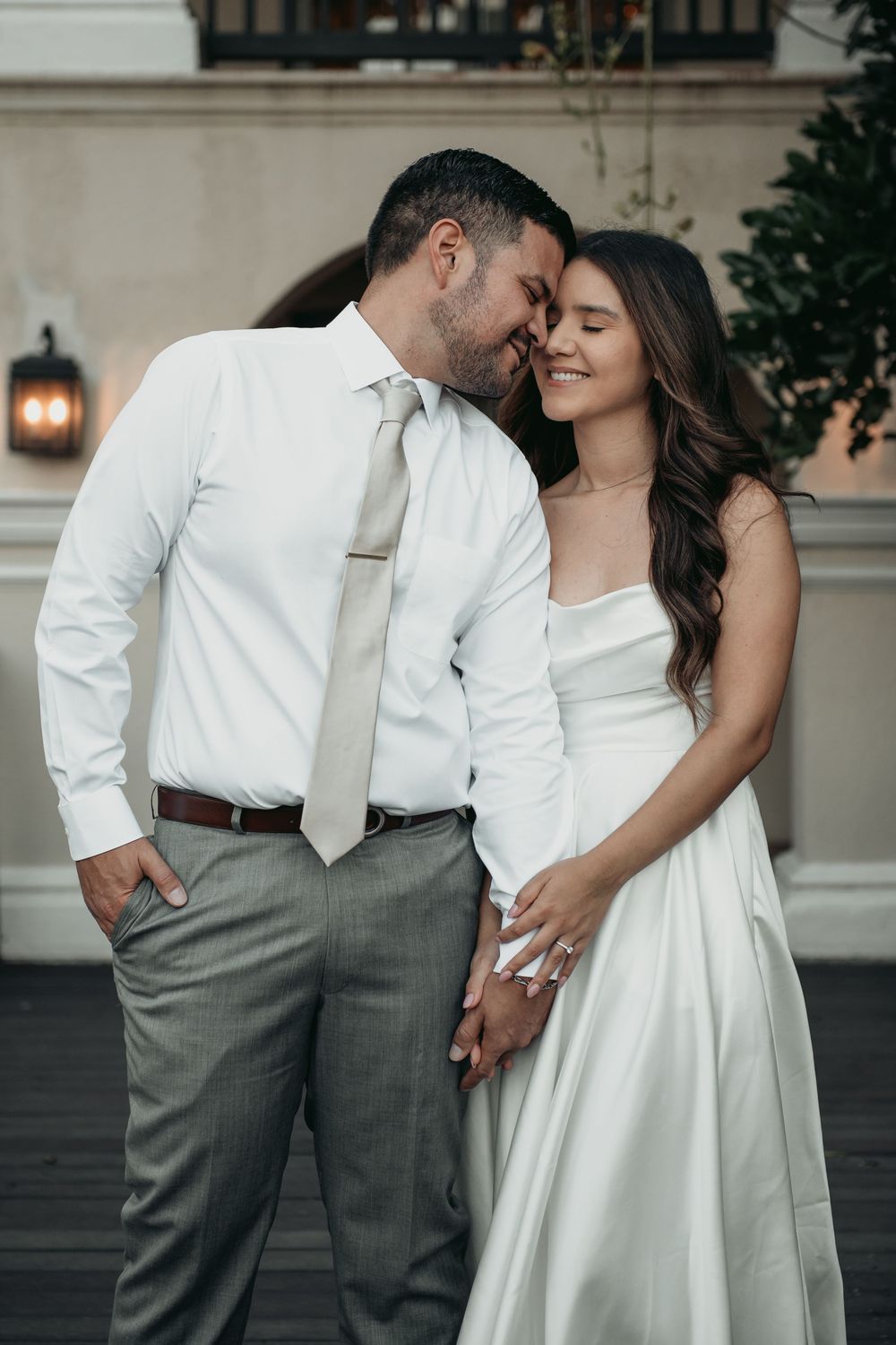 A romantic wedding couple shares an intimate moment outdoors, the bride in white and groom in dress shirt and tie.