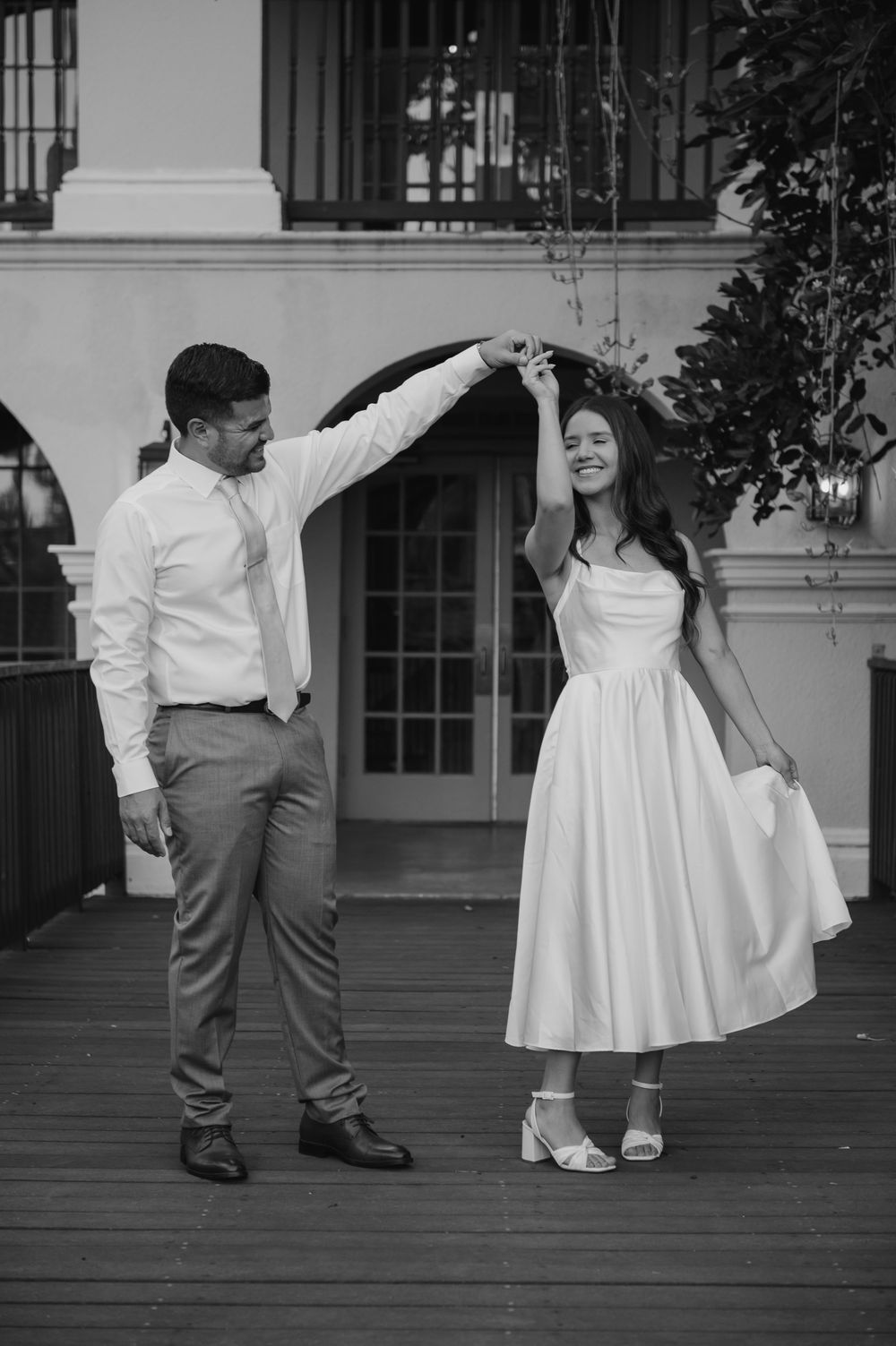 A black and white photo of a couple dancing on a porch, with the woman in a white dress twirling gracefully.