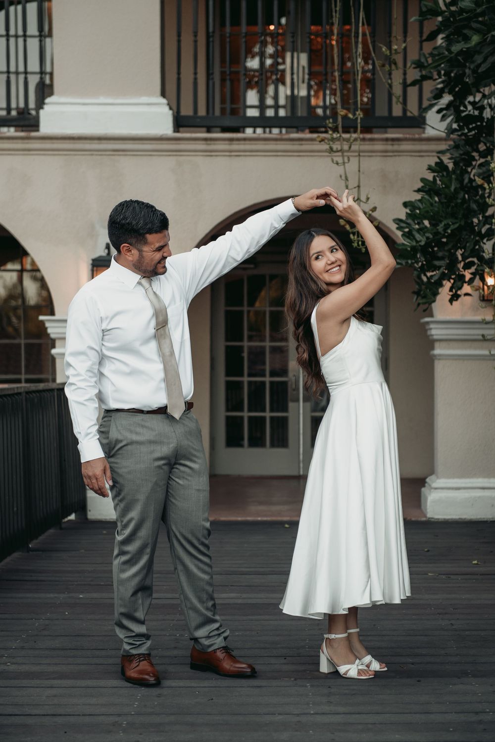 A couple in wedding attire dances together on a dark patio, with the groom in grey pants and white shirt leading a twirl.