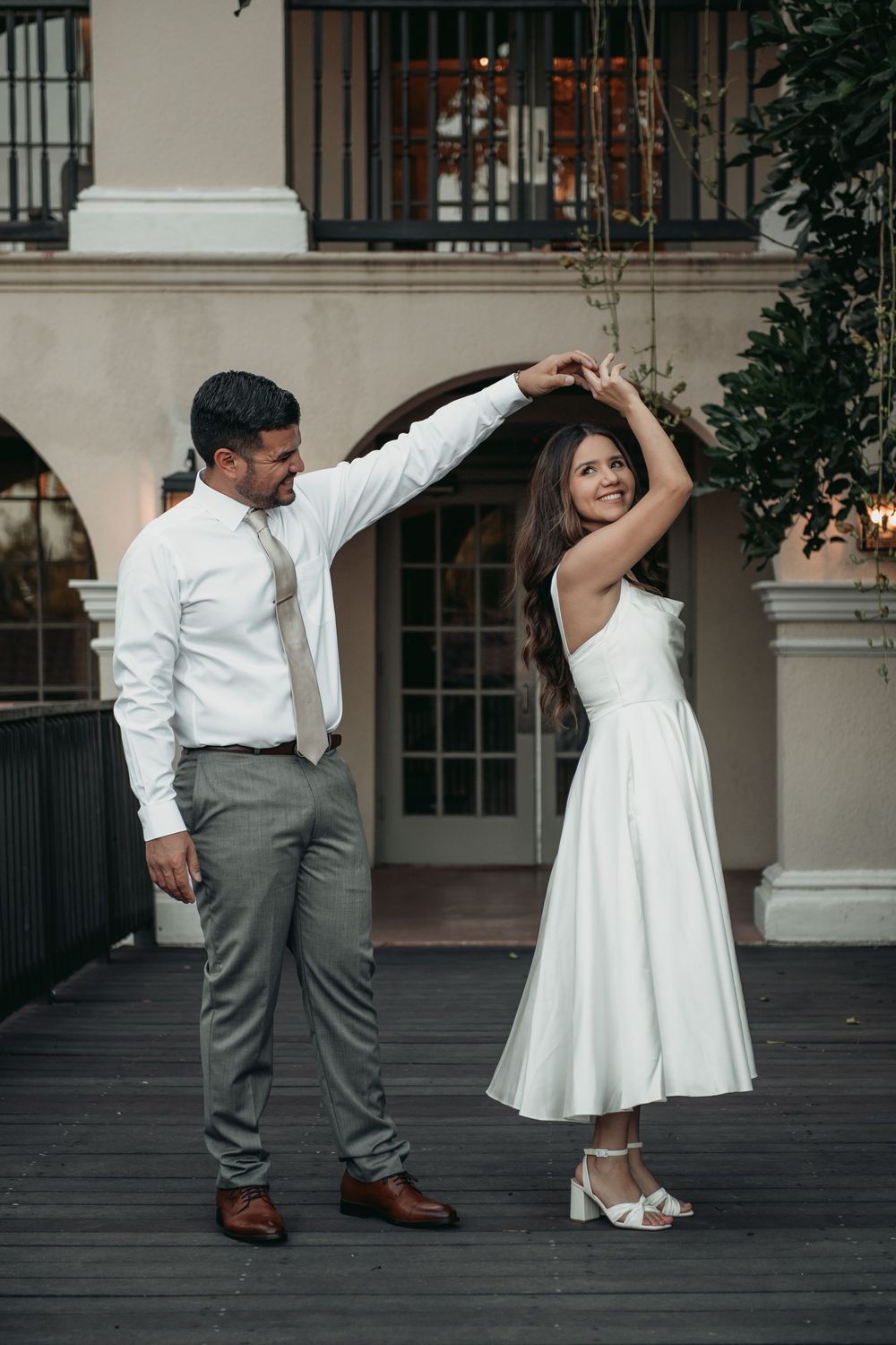 A couple dances together on a dark floor, with the woman in a white dress and man in gray pants and white shirt.