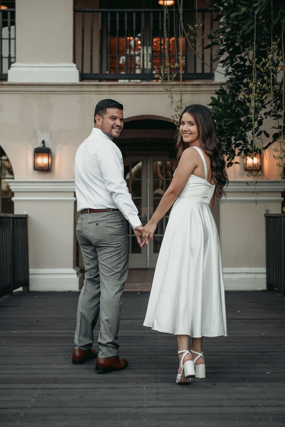 A couple in formal attire holding hands and looking back at the camera on an elegant porch with lanterns.