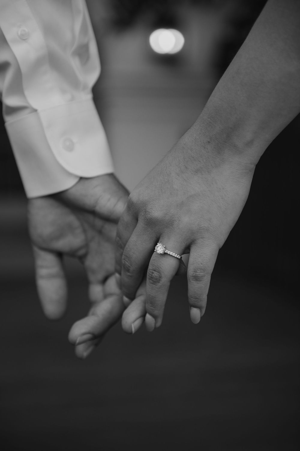 A black and white close-up of two hands clasped together, with an engagement ring visible.
