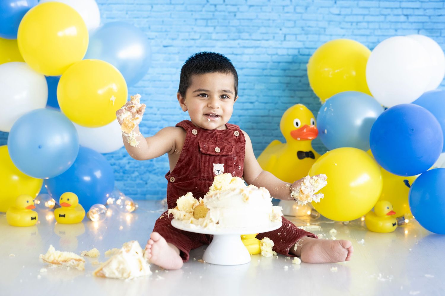 Cake smash photo setup with blue and yellow balloons and rubber duck decorations.