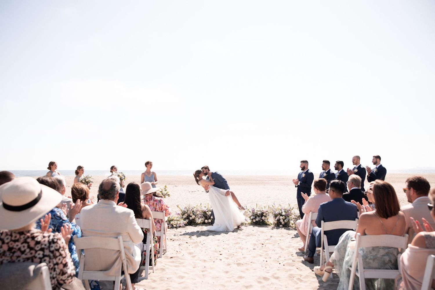 Outdoor wedding ceremony taking place on a coastal terrace with white chairs and wooden arch decorated with flowers.