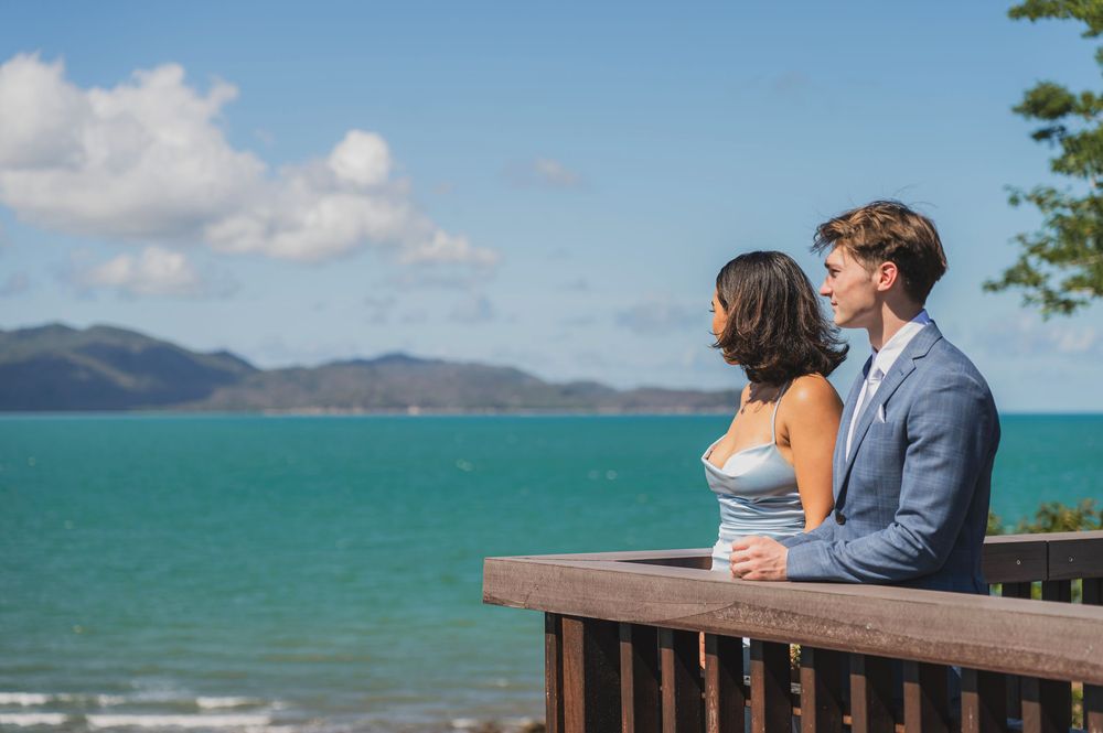 A couple stands at a wooden railing overlooking turquoise waters and mountains on a sunny tropical day.