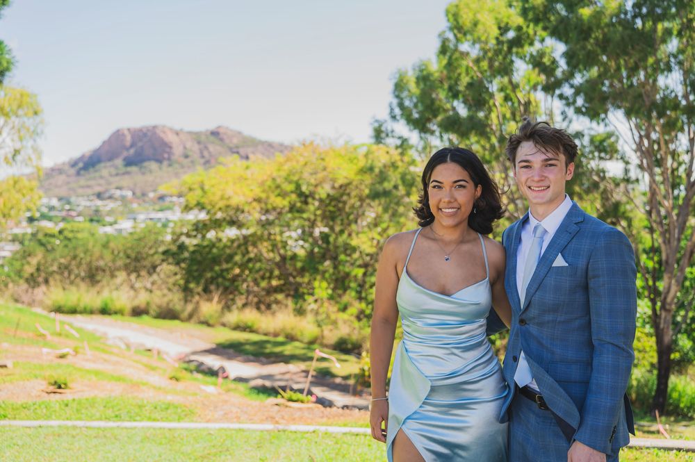 A couple in formal attire poses outdoors in front of scenic mountain landscapes and trees.
